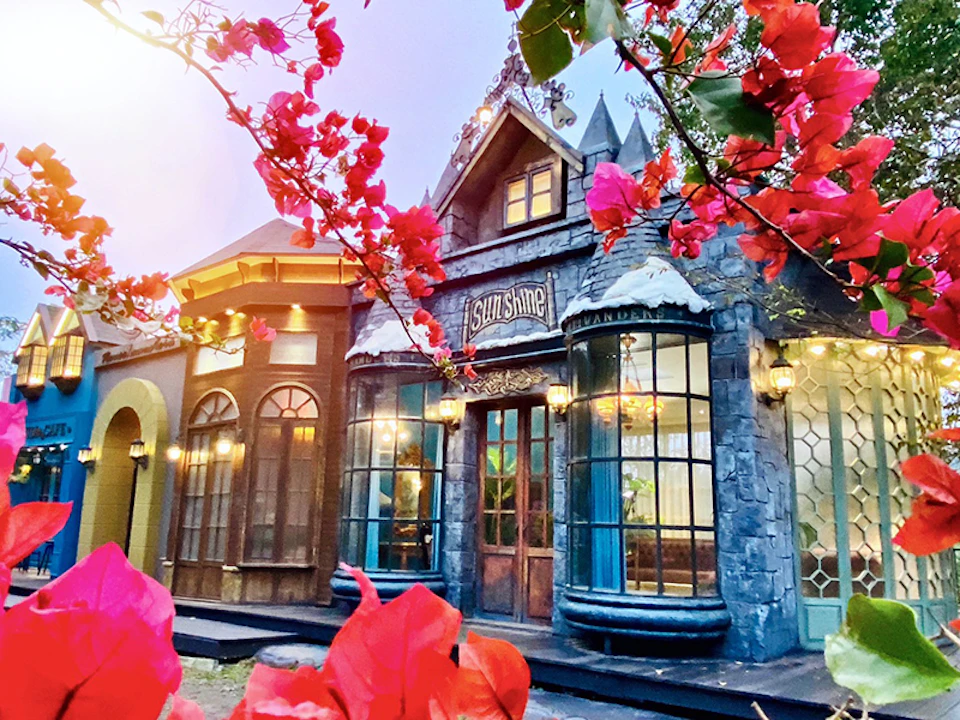 A whimsical cottage-like building with ornate windows and a sign reading "Sunshine," framed by vibrant pink flowers.