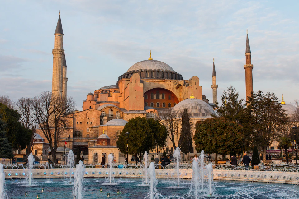 A historic domed building with minarets, trees in the foreground, and a fountain in a plaza under a partly cloudy sky.