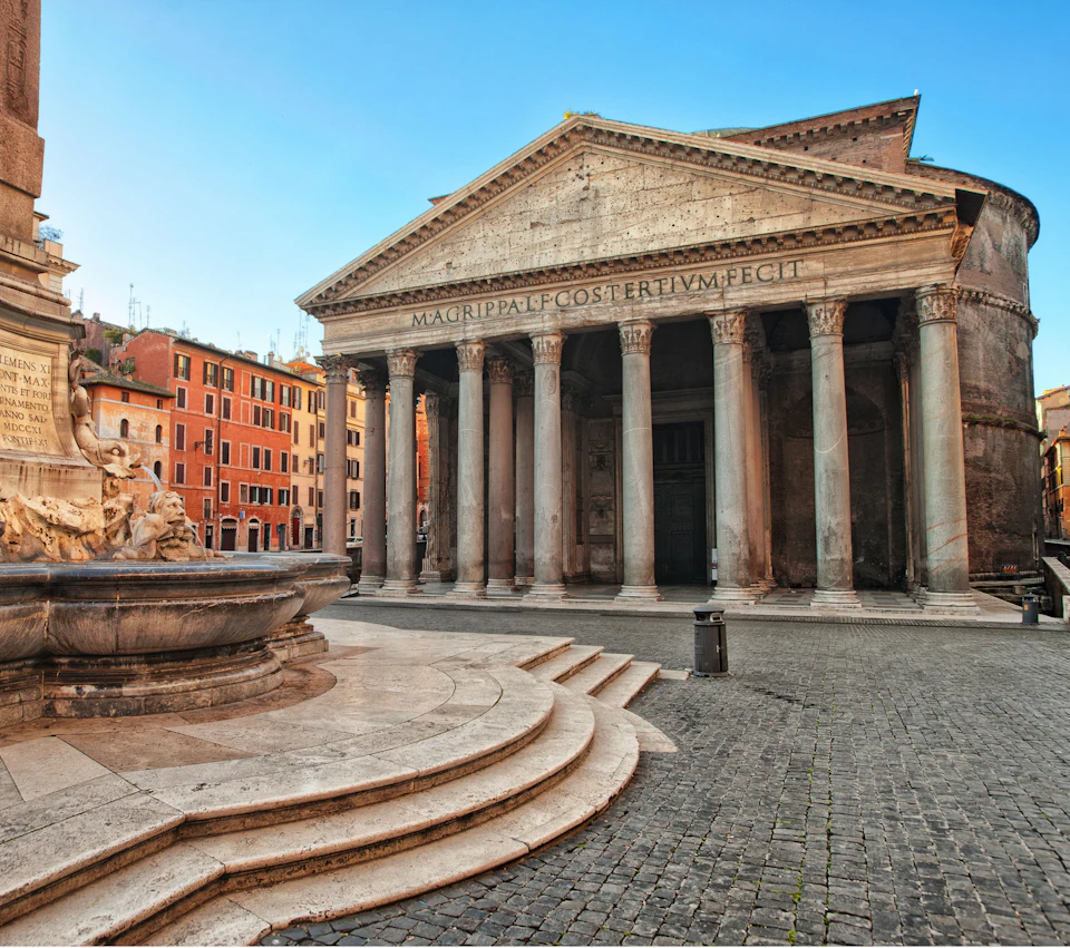 Historic building with tall columns and Latin inscription on the facade, adjacent to a fountain in a cobblestone plaza.