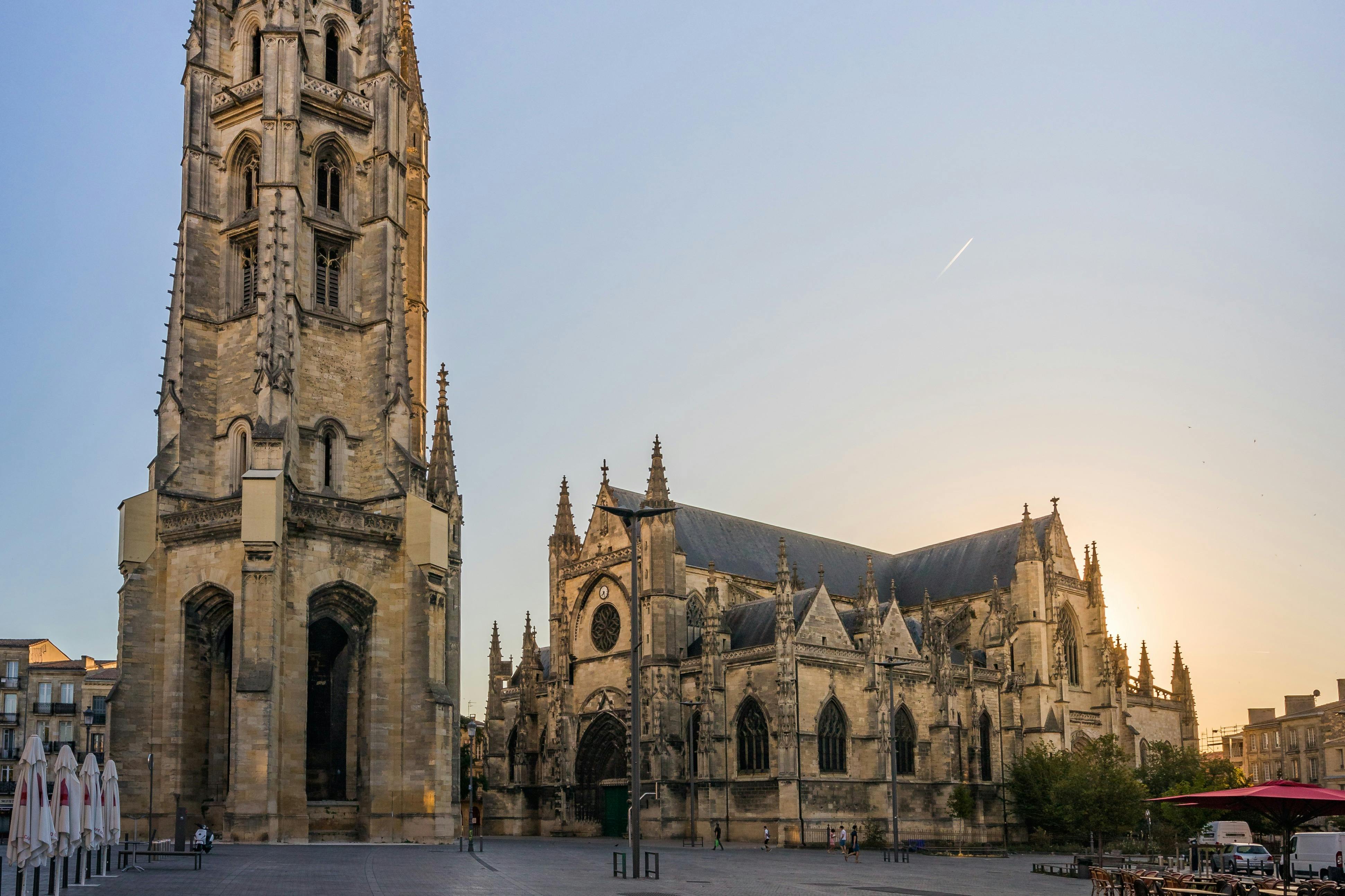 En katedral i gotisk stil med ett högt klocktorn i ett solbelyst torg, omgivet av byggnader och minimal fotgängaraktivitet.