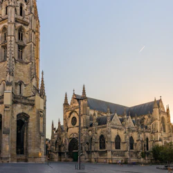 En katedral i gotisk stil med ett högt klocktorn i ett solbelyst torg, omgivet av byggnader och minimal fotgängaraktivitet.