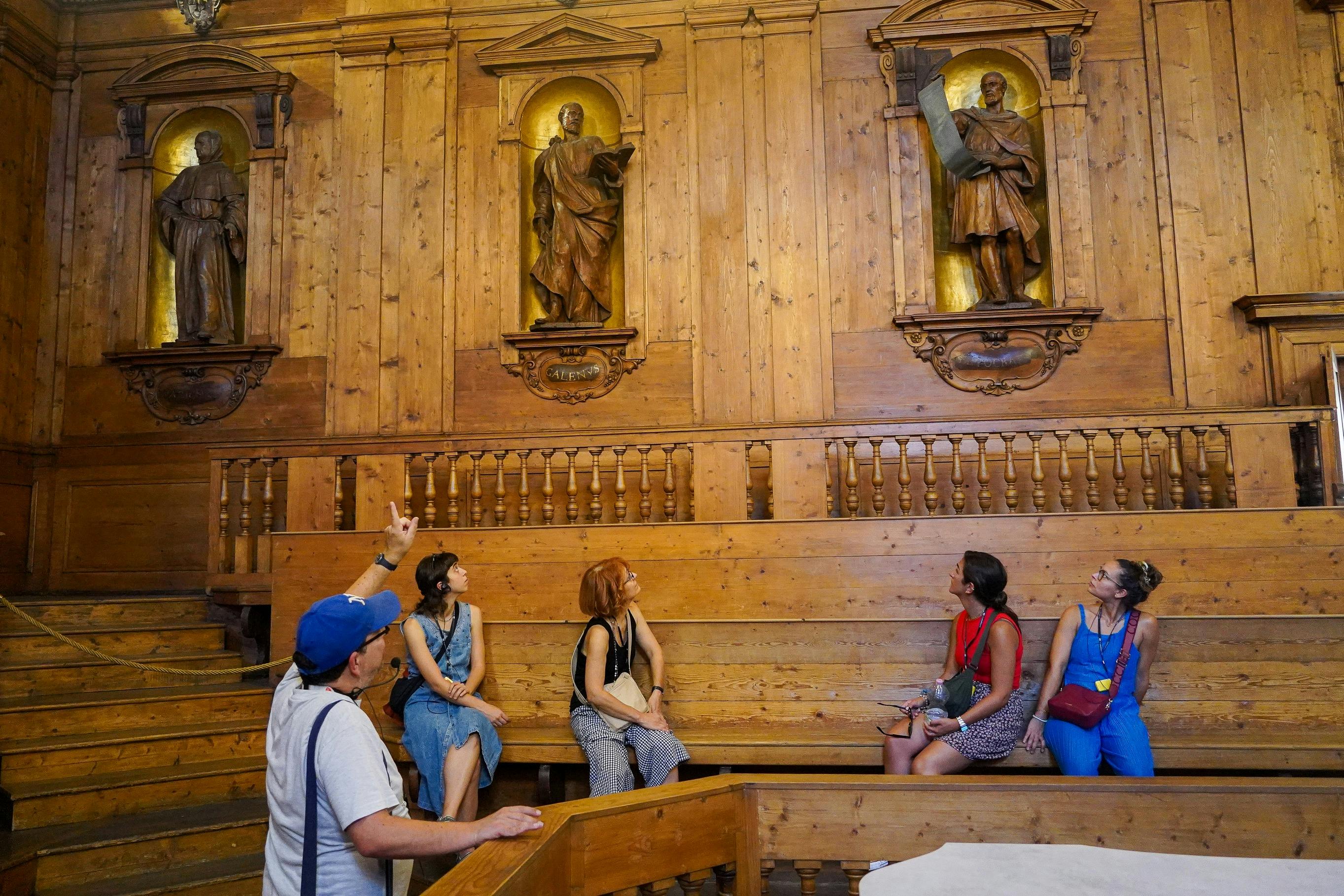 四个人坐在木制长凳上，房间里有木墙和宗教雕像。 导游向雕像示意。