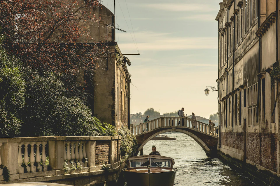 A boat navigates a narrow canal under an old stone bridge, flanked by historic buildings and lush foliage.