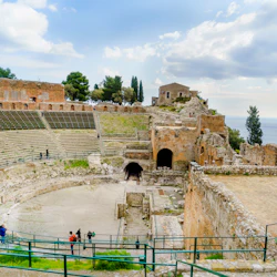 Ancient amphitheater ruins with tiered seating, arched entrances, and scattered visitors, set against a backdrop of trees and cloudy sky.