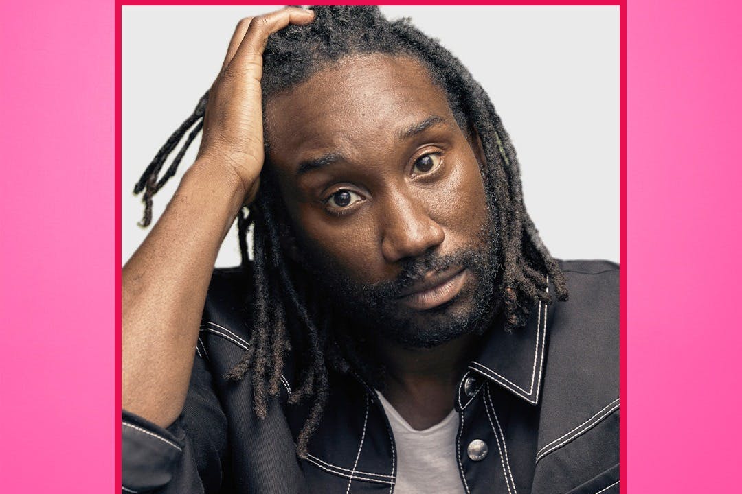 Ένας άντρας με ράστα και γένια, φορώντας ένα σκούρο σακάκι, κρατά το κεφάλι του με το ένα χέρι σε ένα απλό φόντο.