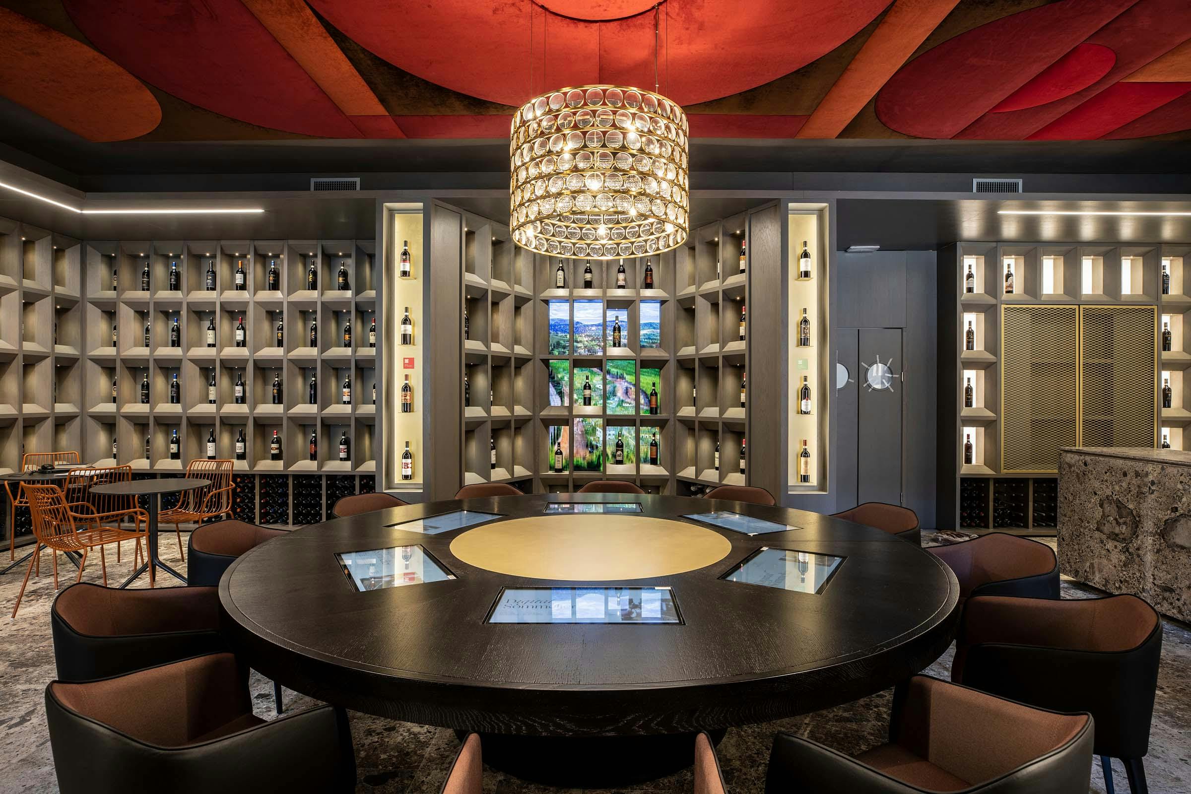 A luxurious room with a circular table, chandelier, and walls lined with shelves displaying bottles and lit photos.