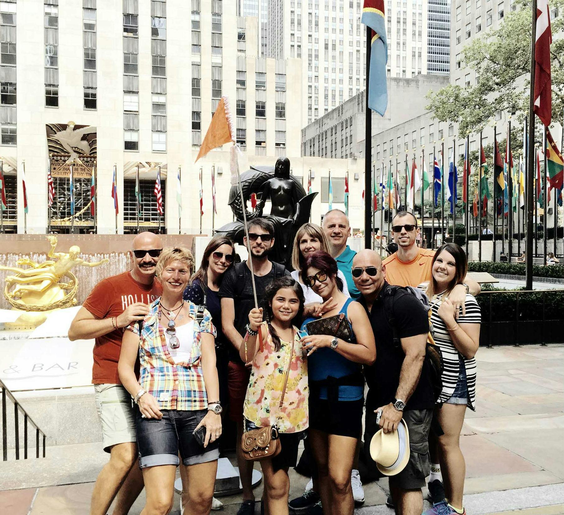 A group of people posing for a photo in front of the Prometheus statue at Rockefeller Center, with numerous flags in the background.