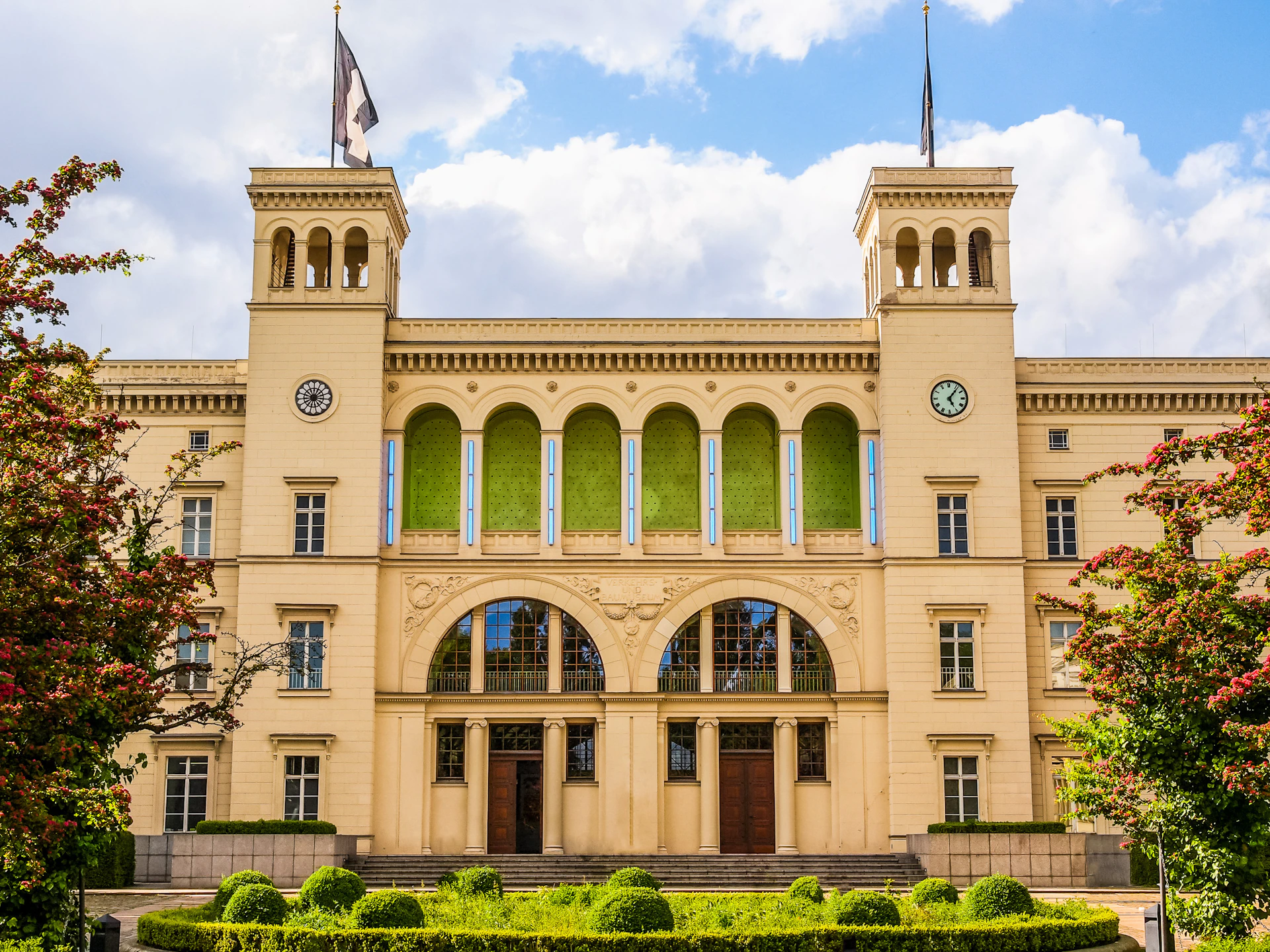 A beige neoclassical building with two towers, multiple arched windows, and two clocks, set against a partly cloudy sky.