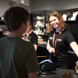 En leende kvinnlig kassörska i en svart skjorta hjälper en kund vid en butiksdisk med plyschdjur och varor i bakgrunden.