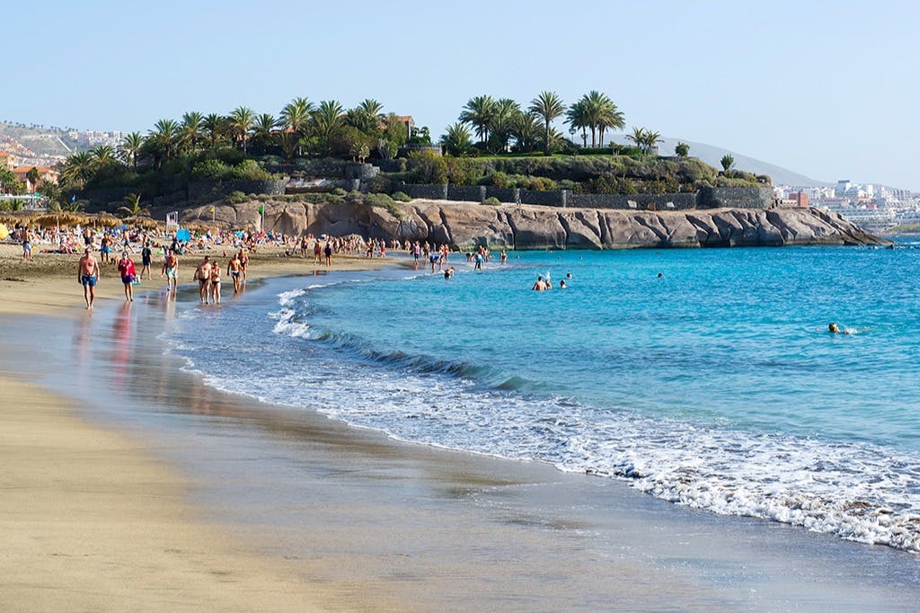 Люди гуляют и купаются на песчаном пляже с прозрачной голубой водой, скалистым побережьем и пальмами на заднем плане.