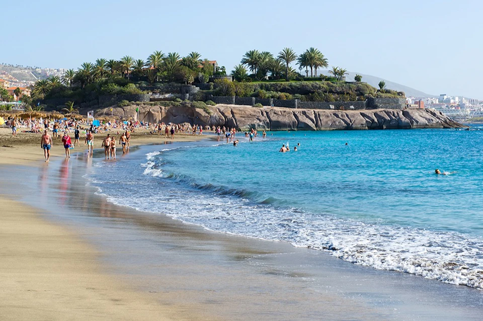 People walking and swimming at a sandy beach with clear blue water, rocky coastline, and palm trees in the background.