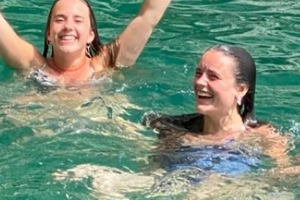 Three people in swimsuits enjoying and raising their arms while swimming in clear turquoise water near rocky cliffs.