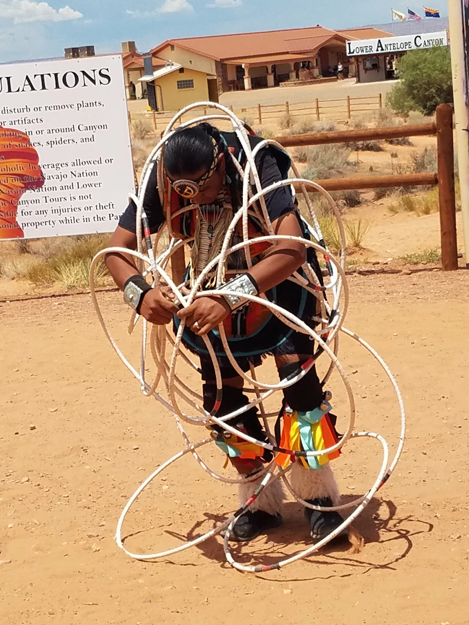 A person dressed in traditional attire performs with multiple hoops in an outdoor setting near a sign and fence.