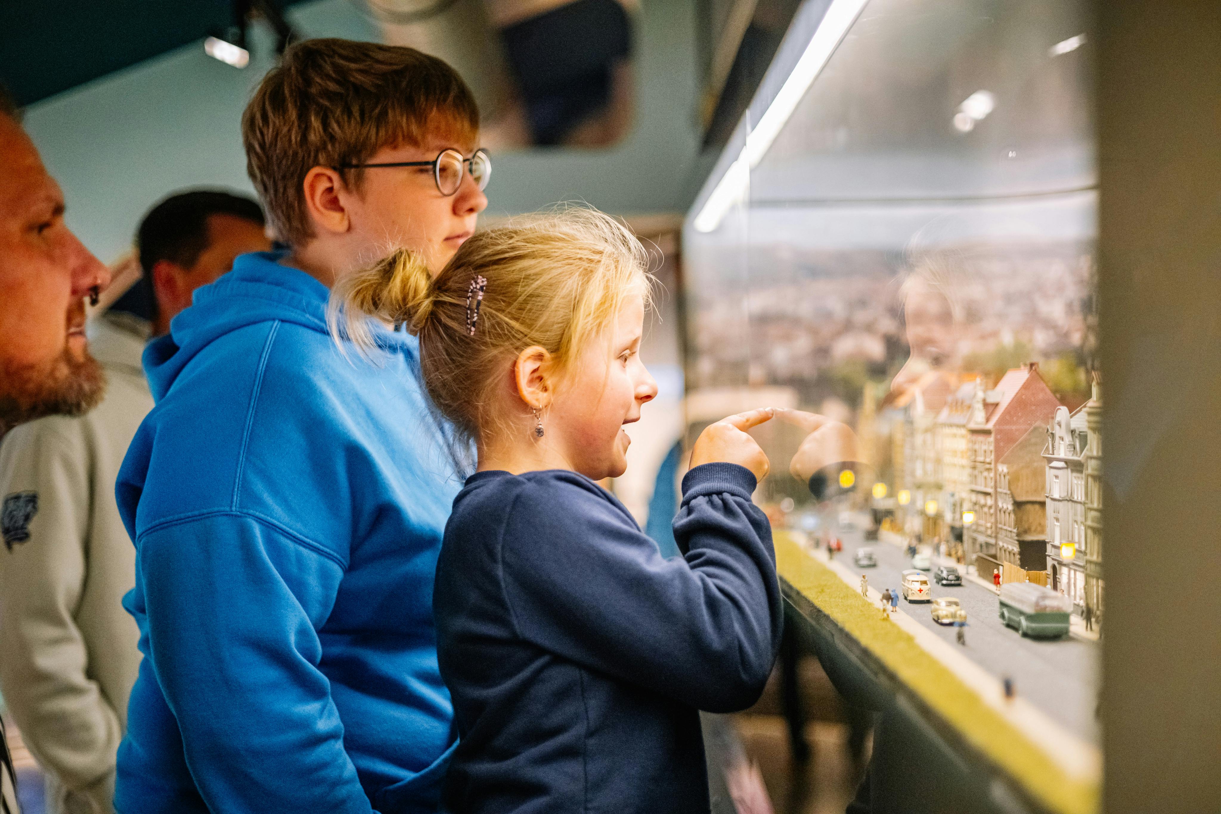 Two children observing a miniature display, one pointing at it, in a well-lit indoor setting.
