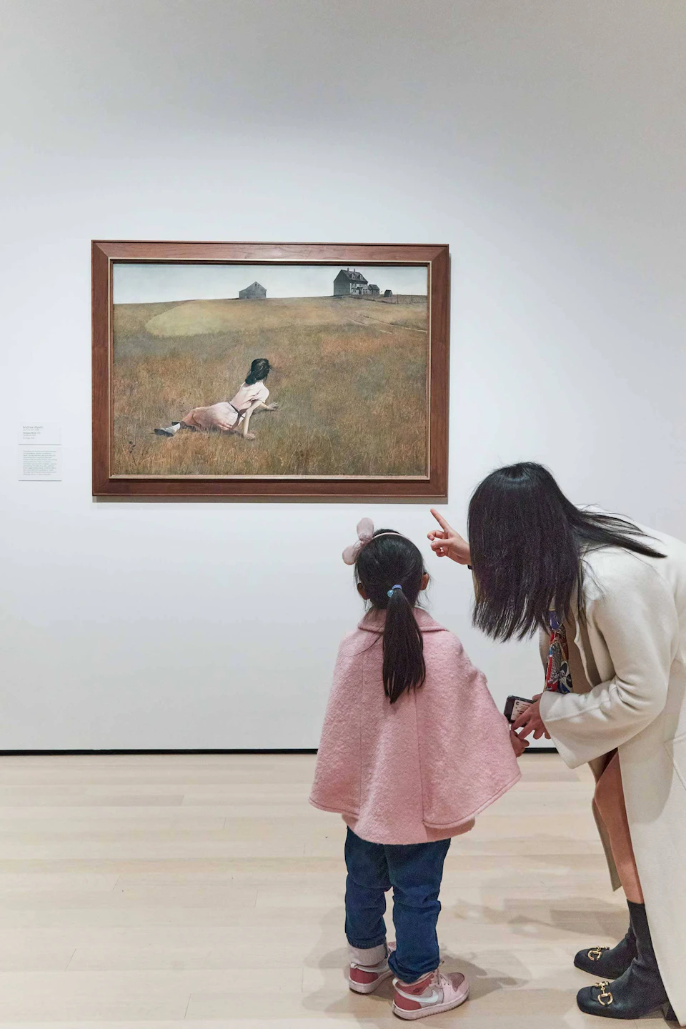 A woman and child in pink clothing view a framed painting of a girl in a field, displayed on a white gallery wall.