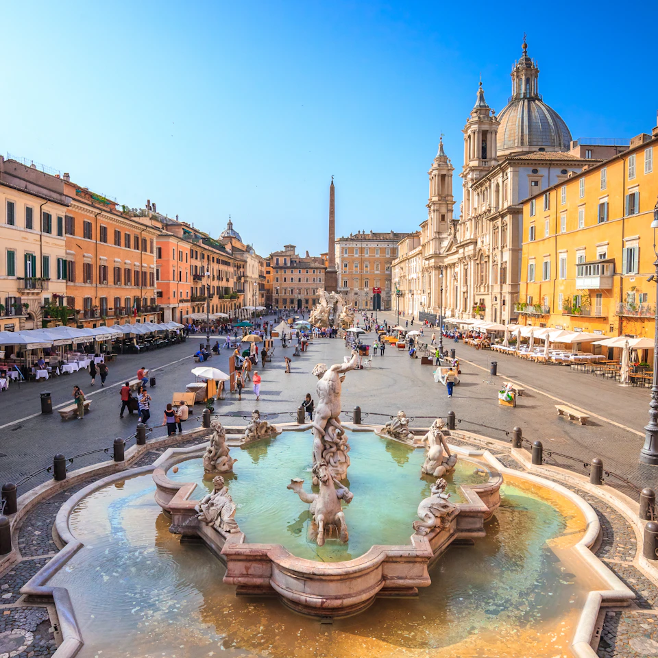 A vibrant city square with a large, ornate fountain, surrounded by historic buildings, outdoor cafes, and people walking or sitting.