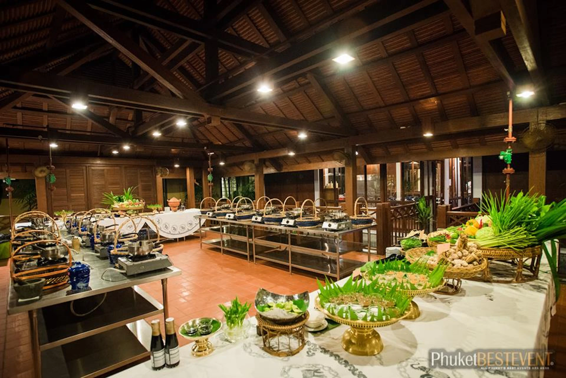 An indoor buffet setup with various dishes displayed under a wooden roof structure, featuring ornate serving platters and green garnishes.