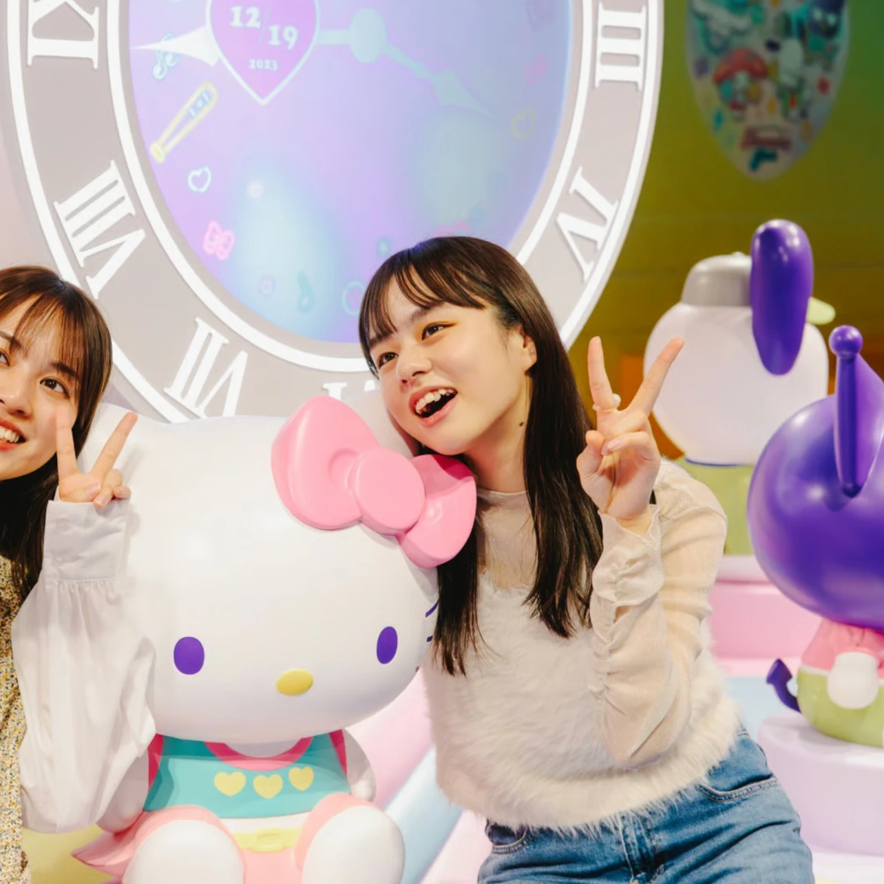 Two people posing with a Hello Kitty statue in a colorful setting with a clock-themed backdrop.