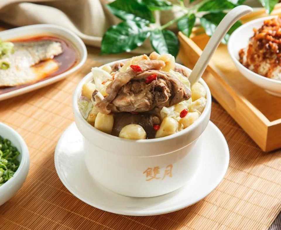 A white bowl with soup containing meat, garlic, and berries, placed on a bamboo mat with a spoon and other dishes nearby.