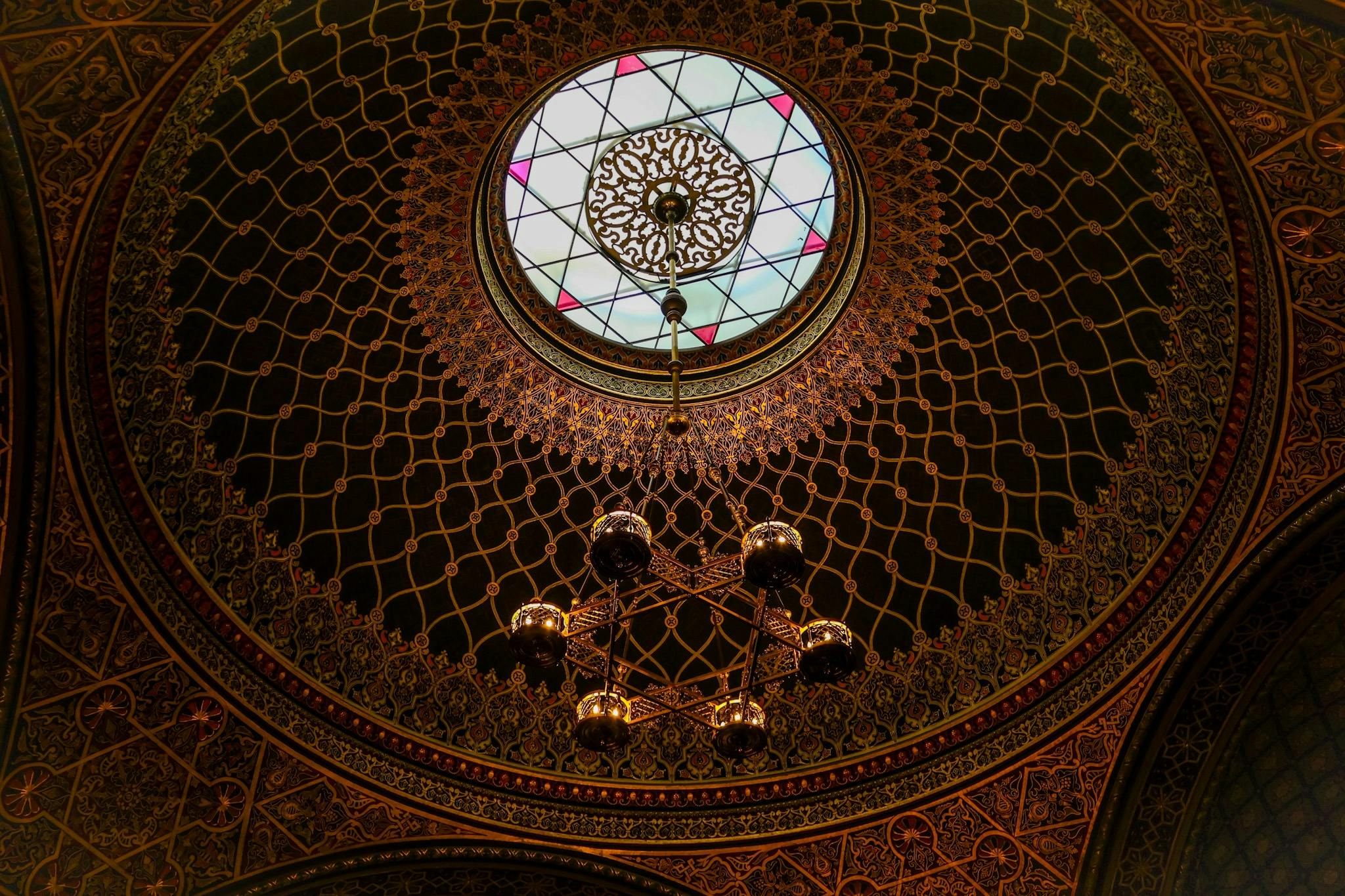 A detailed dome ceiling with intricate patterns, a central stained glass circle, and a decorative chandelier hanging below.