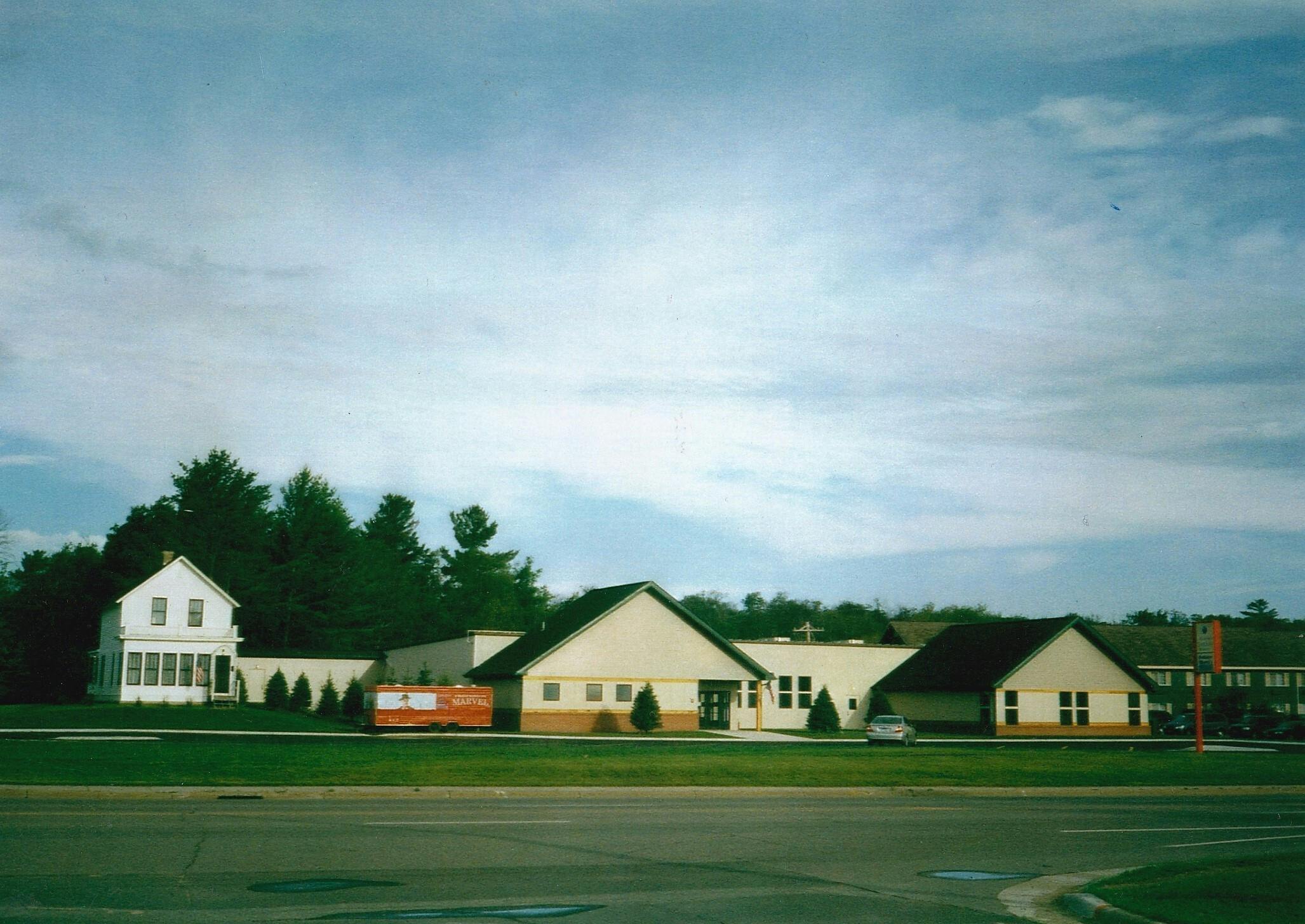 A row of small, modern buildings with triangular roofs, set against a backdrop of trees and a cloudy sky.
