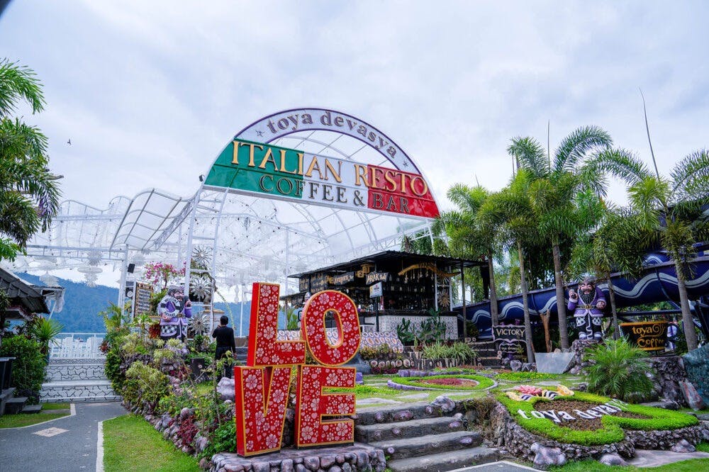 Outdoor Italian restaurant with a large "LOVE" sign, archway, and garden area under a cloudy sky. Palm trees in the background.