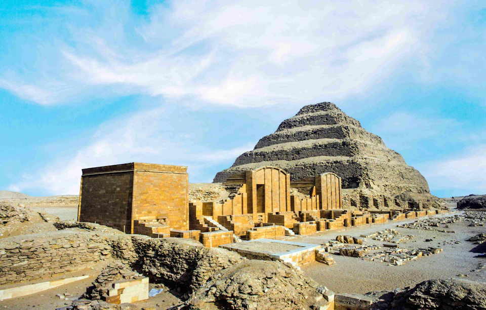 The Step Pyramid of Djoser in Saqqara, Egypt, with surrounding ancient structures under a partly cloudy sky.