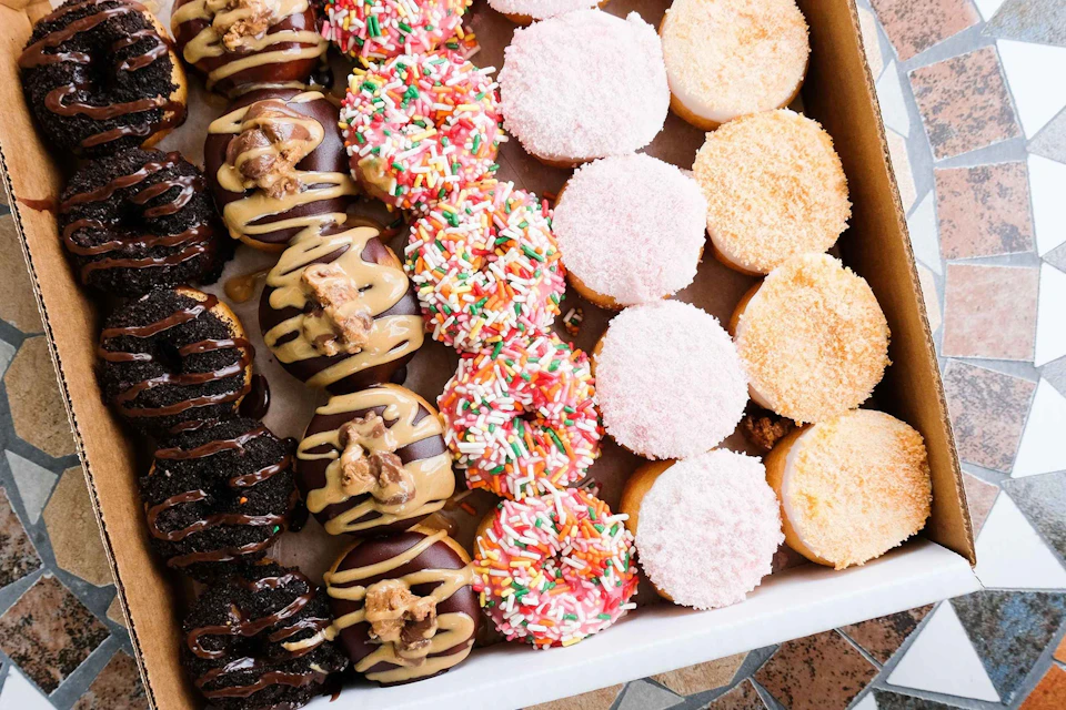 Assorted donuts with various toppings, including sprinkles, glaze, and powdered sugar, arranged in rows in an open box.