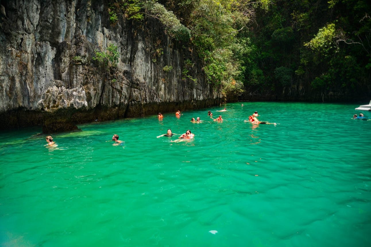 Άτομα που κολυμπούν σε γαλαζοπράσινα νερά κοντά σε βραχώδη βράχο με καταπράσινη βλάστηση.