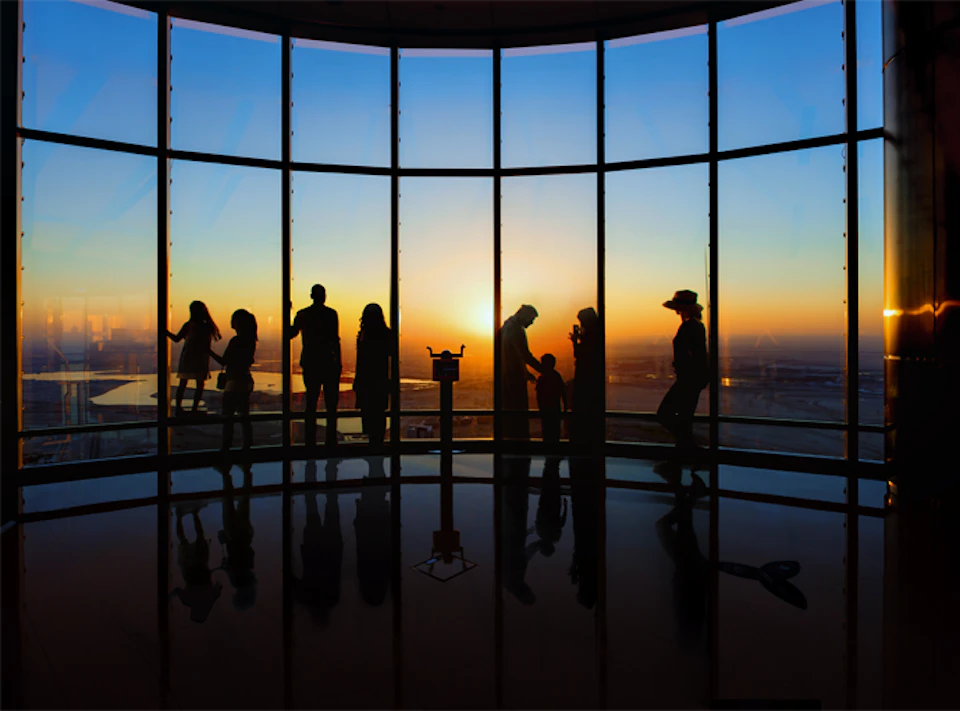 Silhouetted people stand and interact by large windows with a sunset view, overlooking a cityscape.