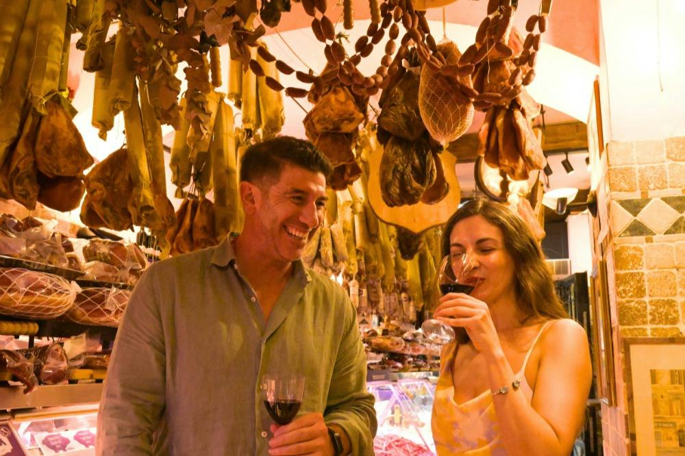 A man and a woman enjoy red wine in a delicatessen with hanging cured meats in the background.