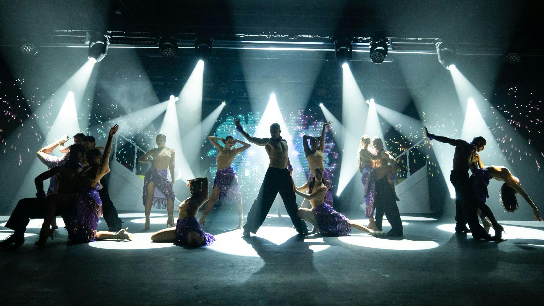 Um grupo de bailarinos em poses dramáticas sob a luz brilhante do palco, vestindo roupas coordenadas com detalhes em roxo.