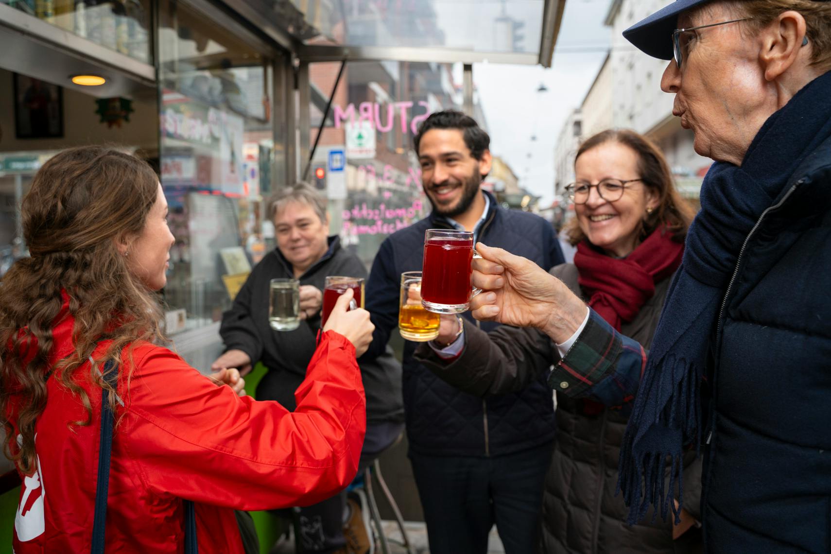 Um grupo de pessoas ao ar livre, sorrindo e segurando copos de bebidas quentes, parecendo brindar numa atmosfera alegre.