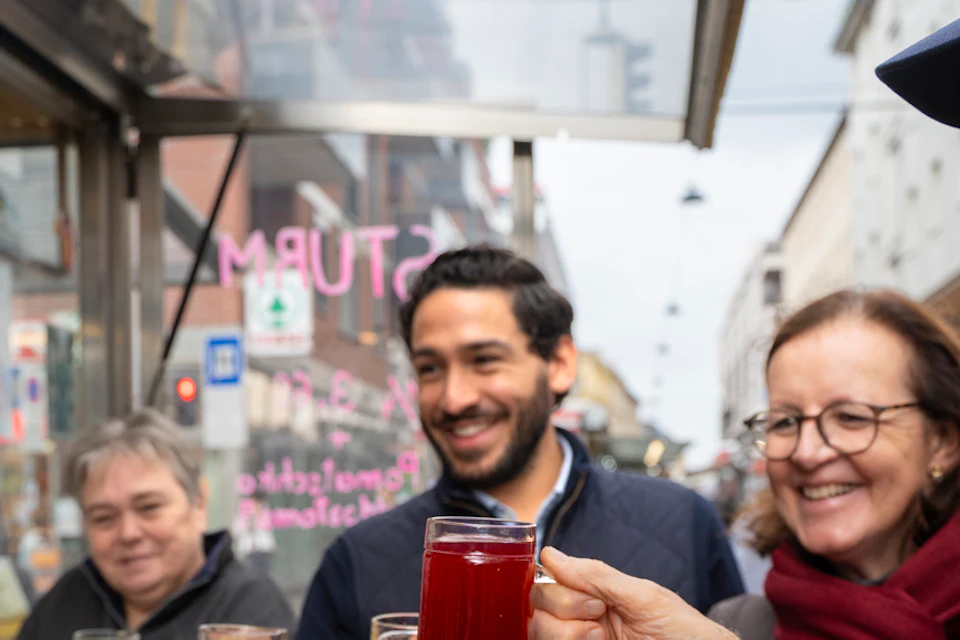 A group of people outdoors smiling and holding glasses of hot beverages, appearing to toast in a cheerful atmosphere.