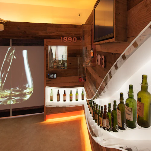 Room with a wooden interior, a row of whiskey bottles on a curved shelf, a projection of liquid pouring into a glass, and "1990" and "2013" wall signs.