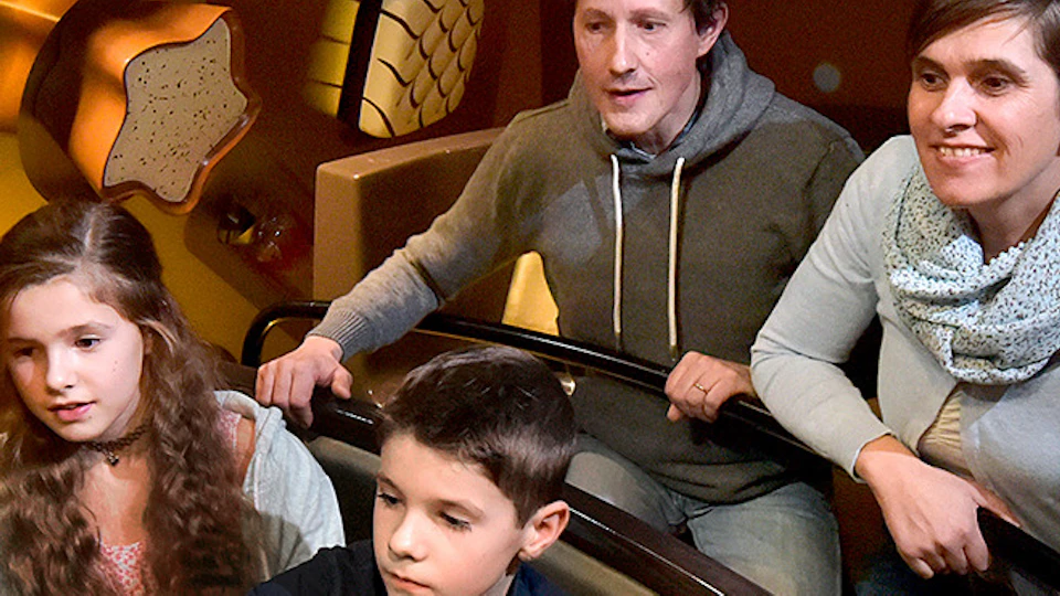 Family of four on a dark ride attraction, with walls decorated with large heart-shaped and other chocolate molds.