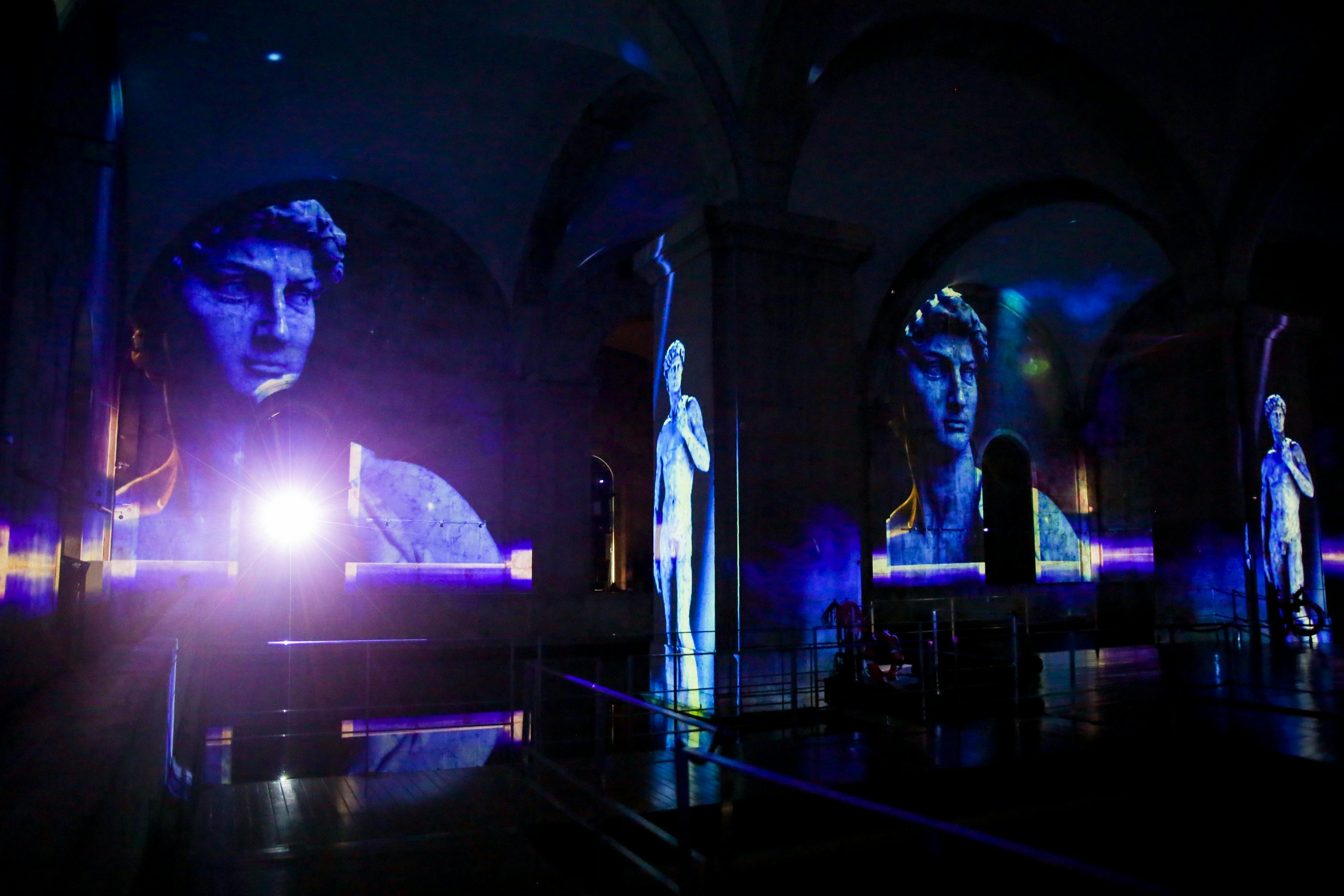 アーチ型の壁に照らされたミケランジェロのダビデ像の大きな投影画像と、明るいフレアを生み出すスポットライトが付いた薄暗い部屋。