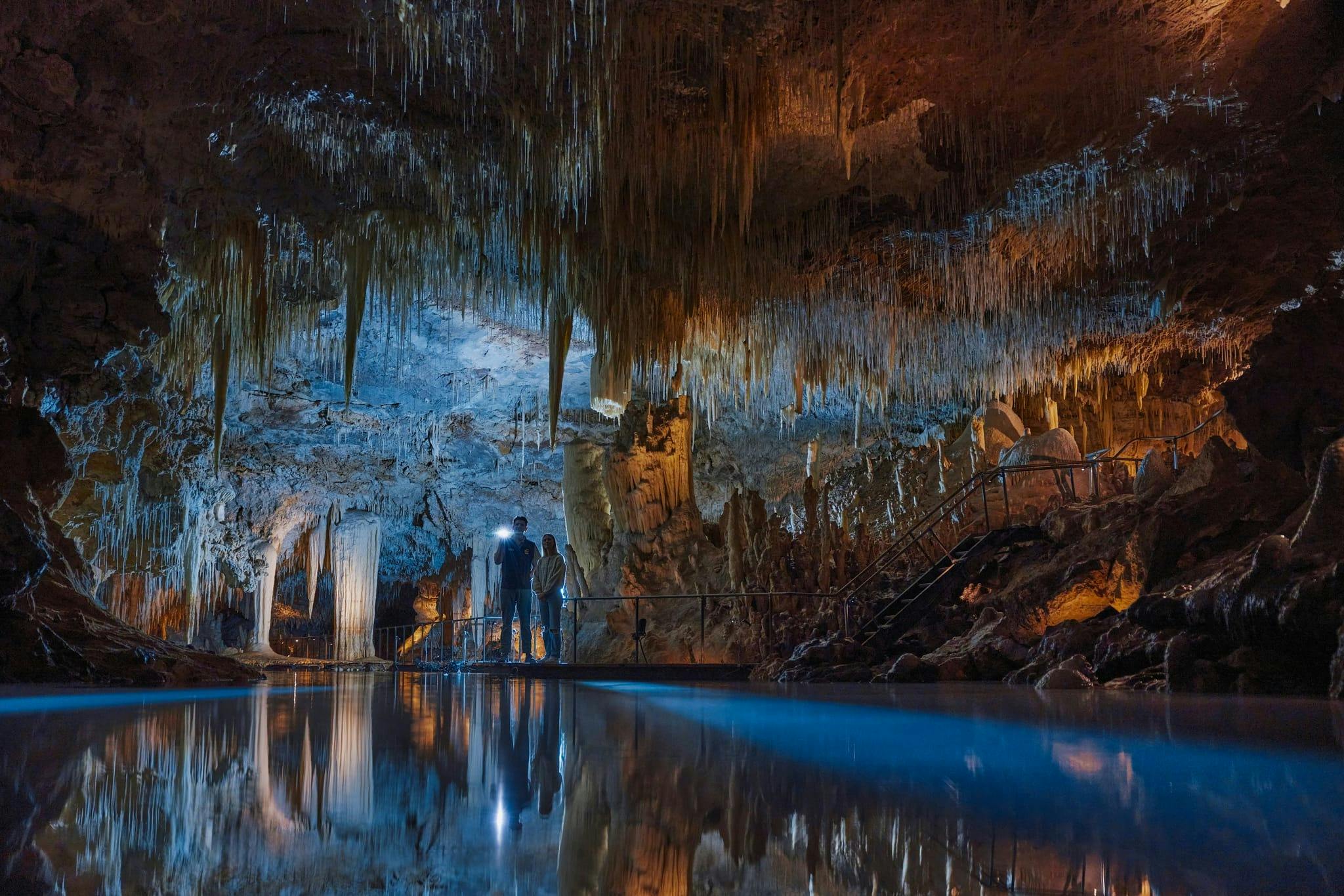 Two people stand on a platform in a cave with stalactites and reflected blue water. One holds a flashlight.