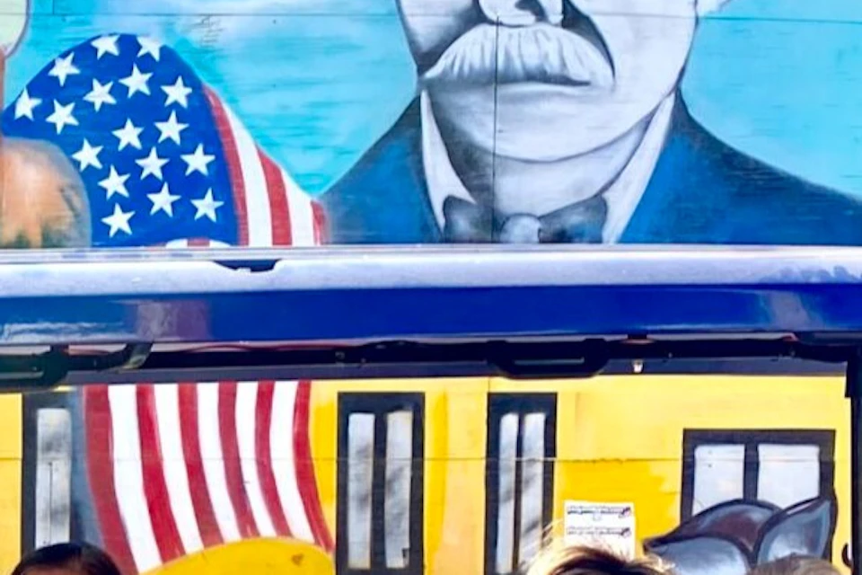 Three women sitting in a blue golf cart in front of a colorful mural with buildings, flags, and a portrait on a brick wall.
