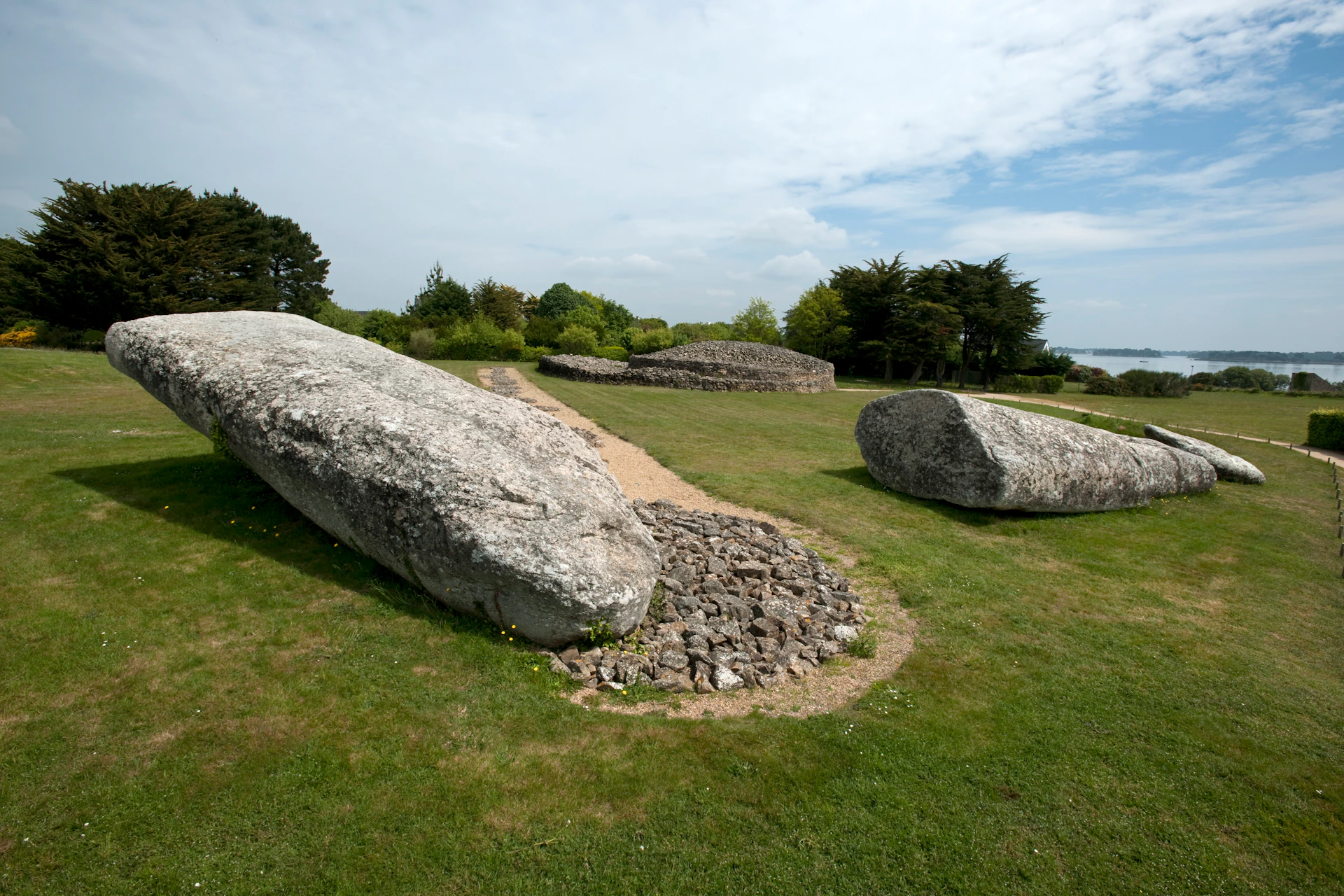 Large ancient stones surrounded by grass and a gravel path, with trees and a cloudy sky in the background.