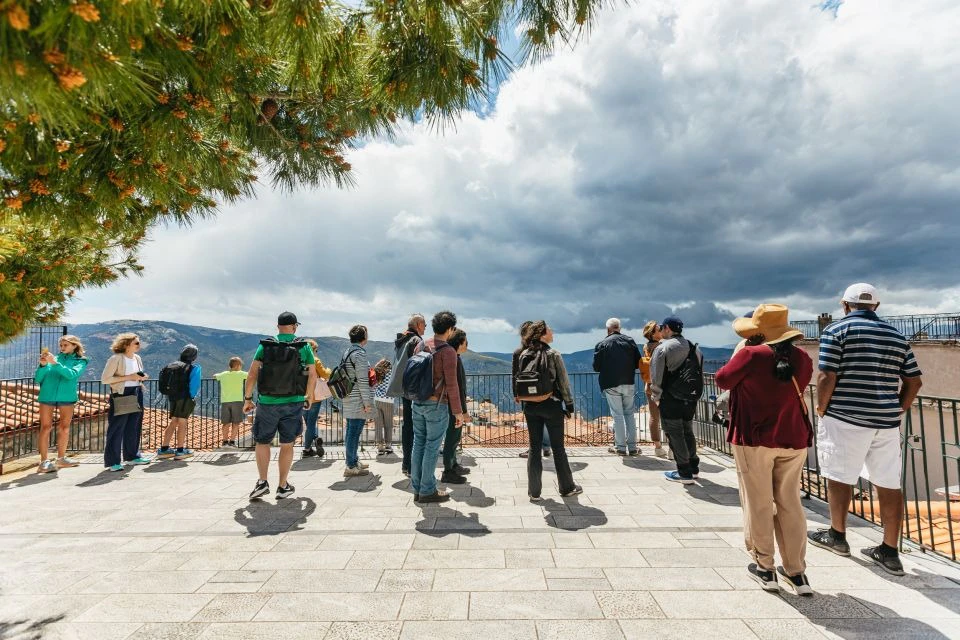 A group of people standing on a tiled lookout platform, under a cloudy sky, with a tree overhead and mountains in the background.