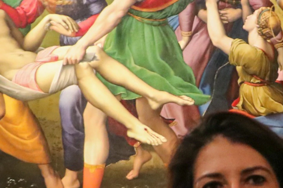 A woman gestures while speaking in front of a large framed painting in a museum, with other visitors in the background.