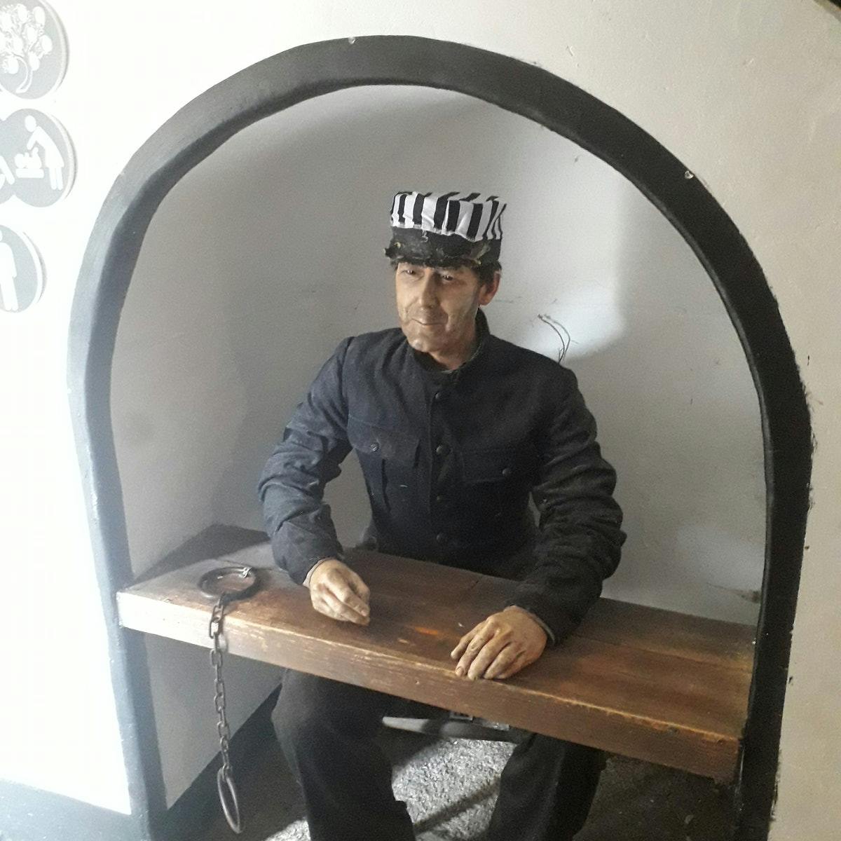 Figurína oblečená jako vězeň v černé uniformě a pruhovaném klobouku, sedící za dřevěným pultem s připevněnou pouty.