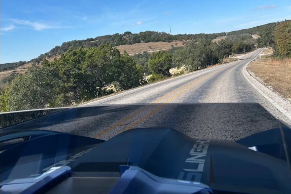 A rural road surrounded by trees and hills under a blue sky, viewed from a vehicle with part of its dashboard in the foreground.