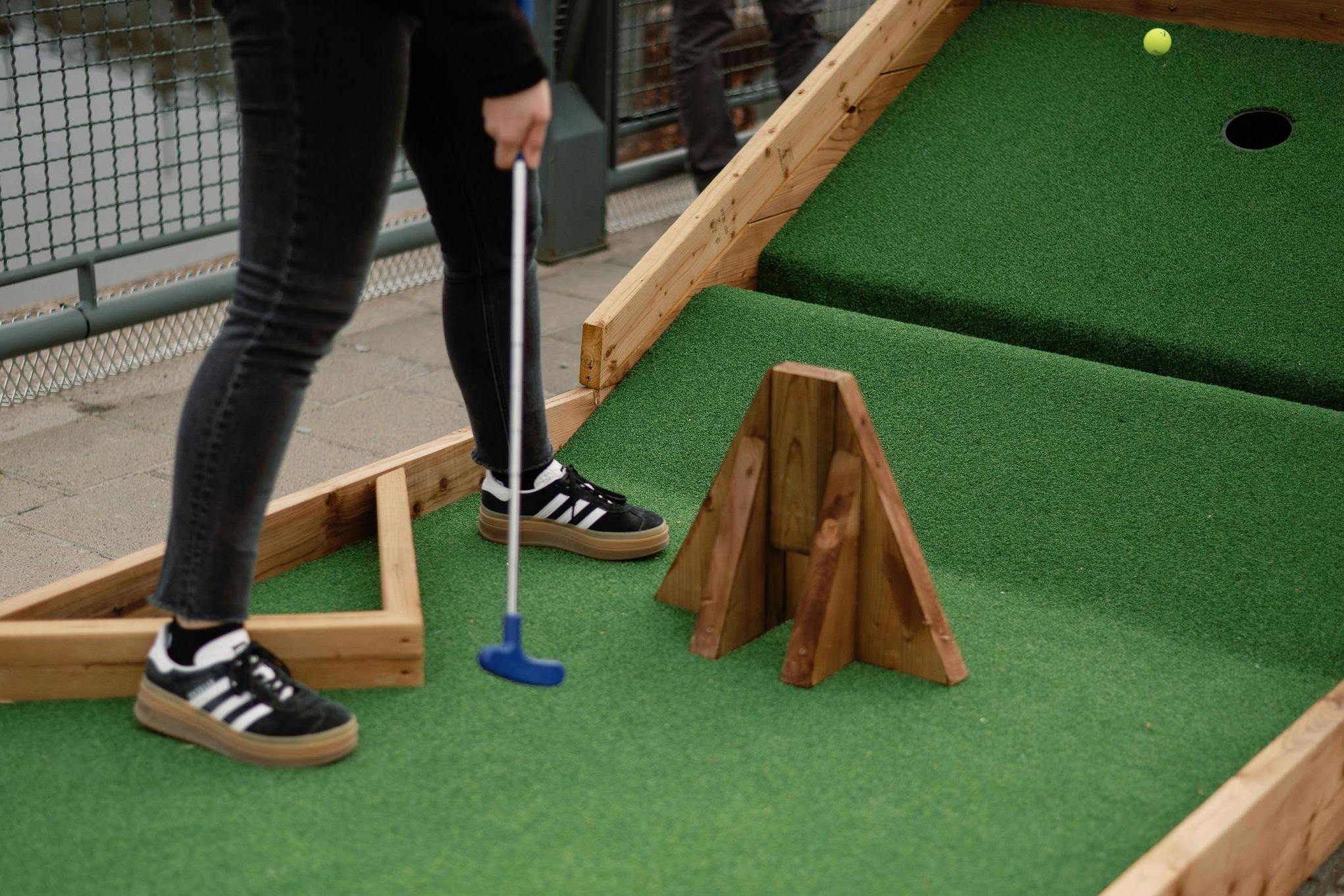 Uma pessoa a jogar mini-golfe, segurando um taco azul perto de um obstáculo de madeira num campo de relva artificial verde.