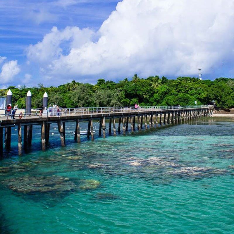 A long pier stretches over clear, turquoise water with people walking on it, leading to lush green trees under a partly cloudy sky.