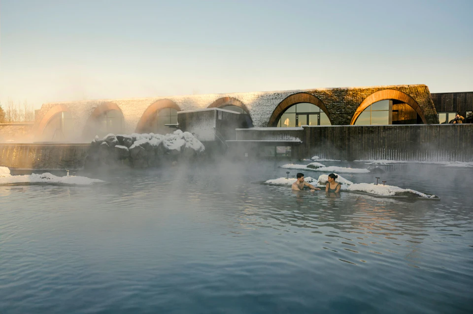 Two people sit in a steaming outdoor hot spring pool near a snow-covered building with arched windows under a clear sky.