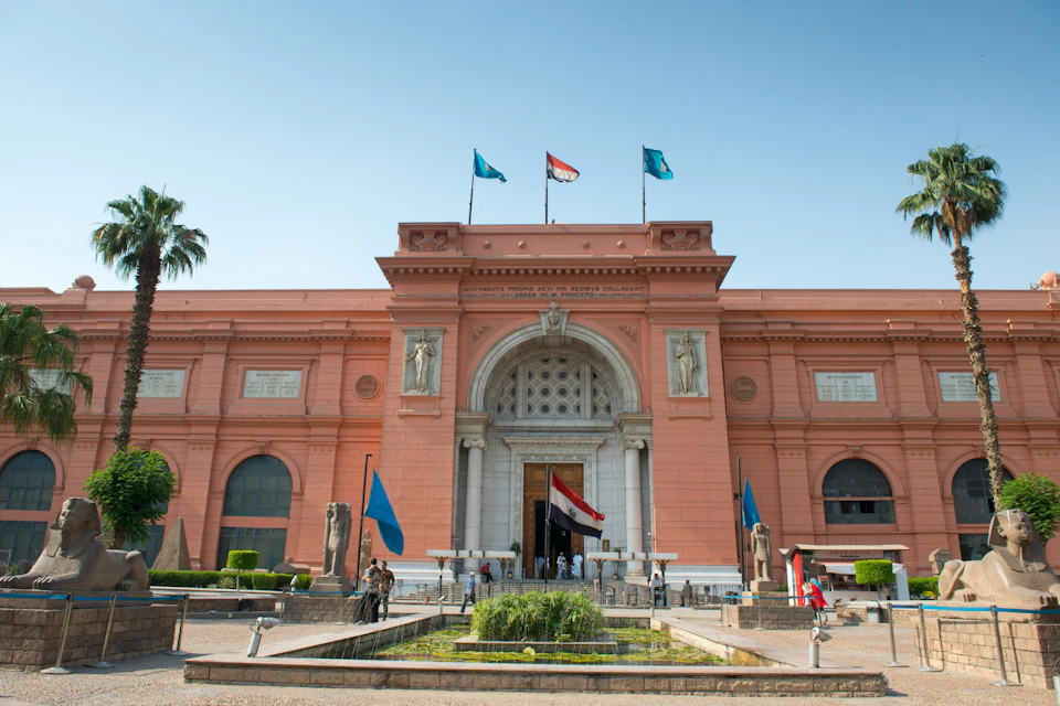 A large red building with an arched entrance, three flags on top, statues on either side, and a fountain in the foreground.