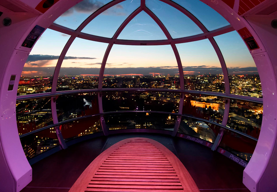 Interior of a Ferris wheel capsule overlooking a cityscape at dusk, with illuminated buildings and a red bench in the foreground.