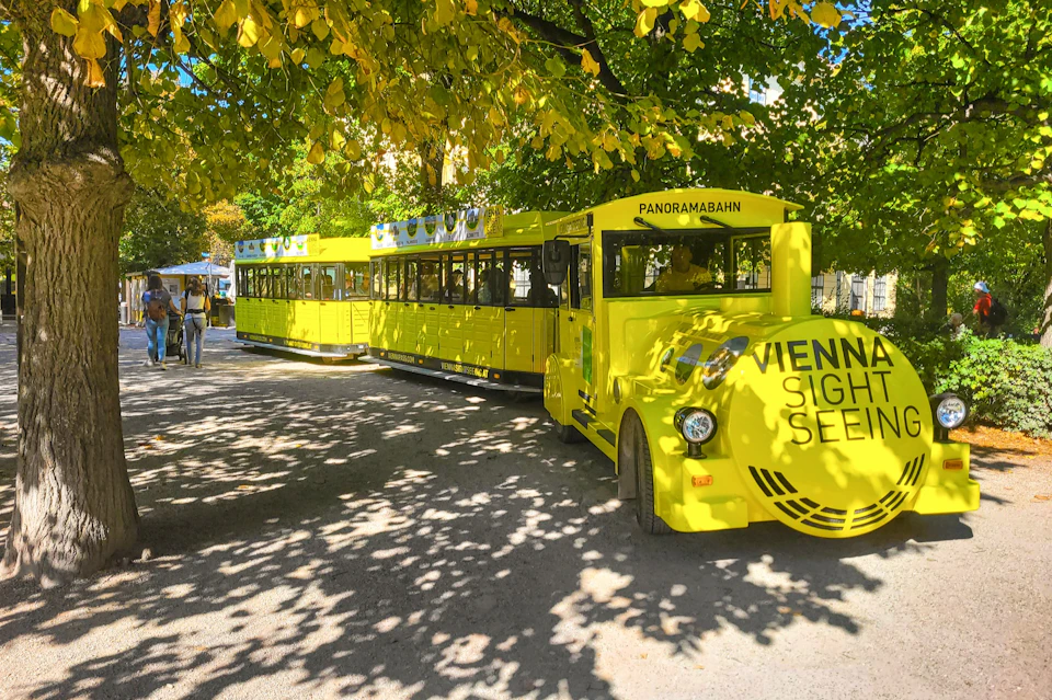 A bright yellow sightseeing train labeled "Vienna Sightseeing" parked under trees with people walking nearby.